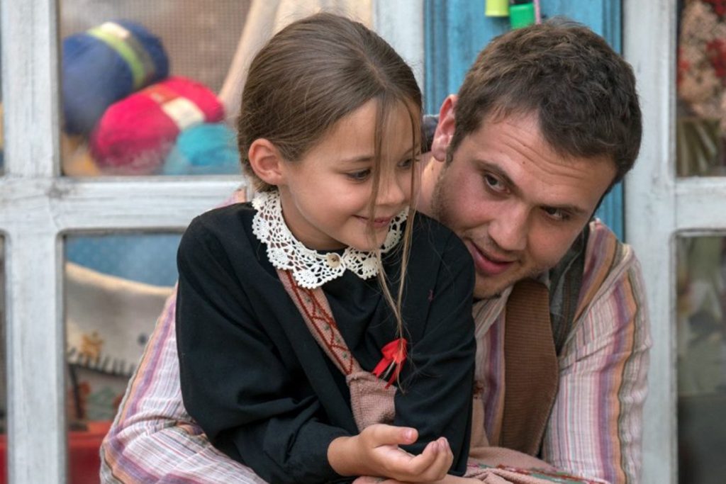 as personagens do filme Milagre na Cela 7, Memo e Ova, estão sentados em frente a uma loja. Ambos estão sorrindo e felizes.