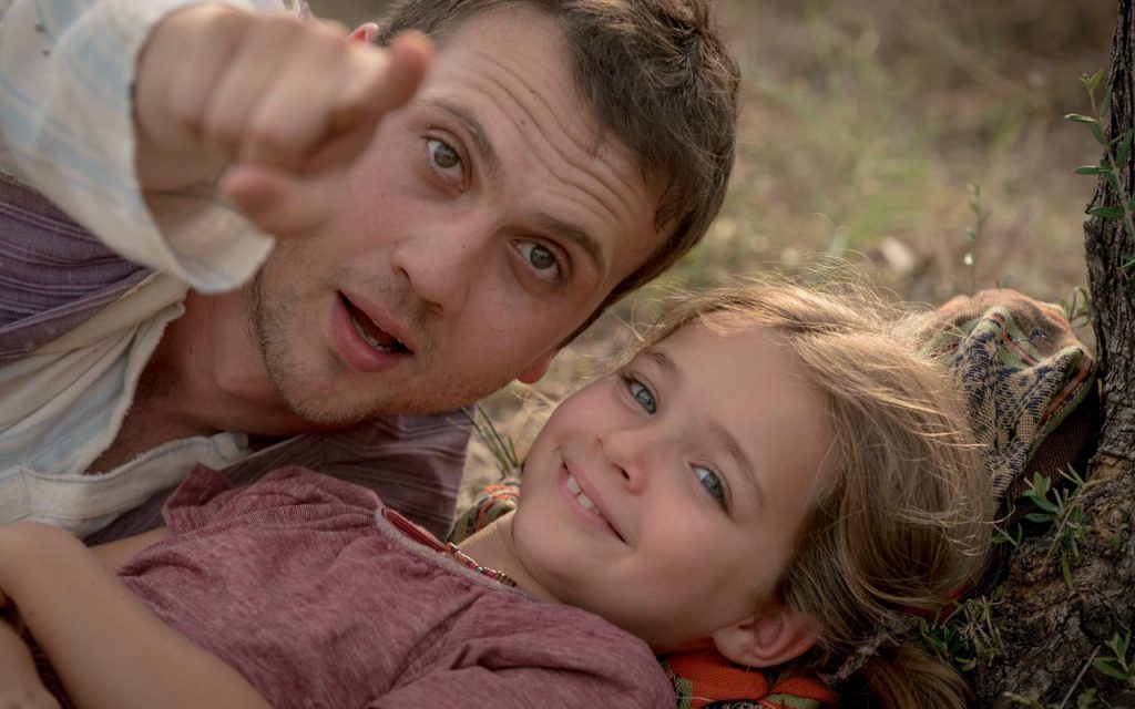 Os atores principais do filme Milagre na Cela 7 estão deitados na grama. Ova está sorrindo, enquanto Memo está mostrando alguma coisa para a menina