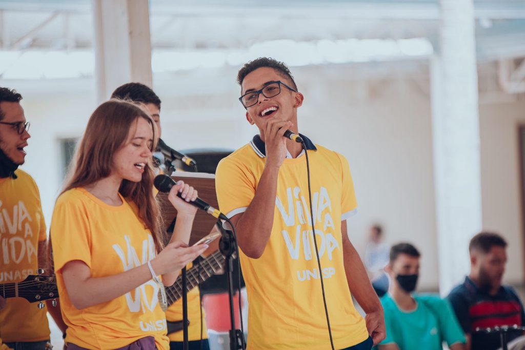 Dois jovens de camiseta amarela cantando para os colegas no pátio do colégio.